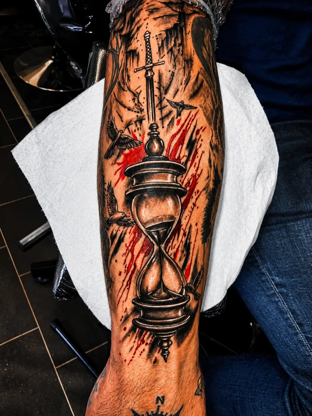Trash polka style forearm tattoo of an hourglass pierced by a sword with red paint splashes and birds, black and red realism at N9 Tattoo, Edmonton, North London